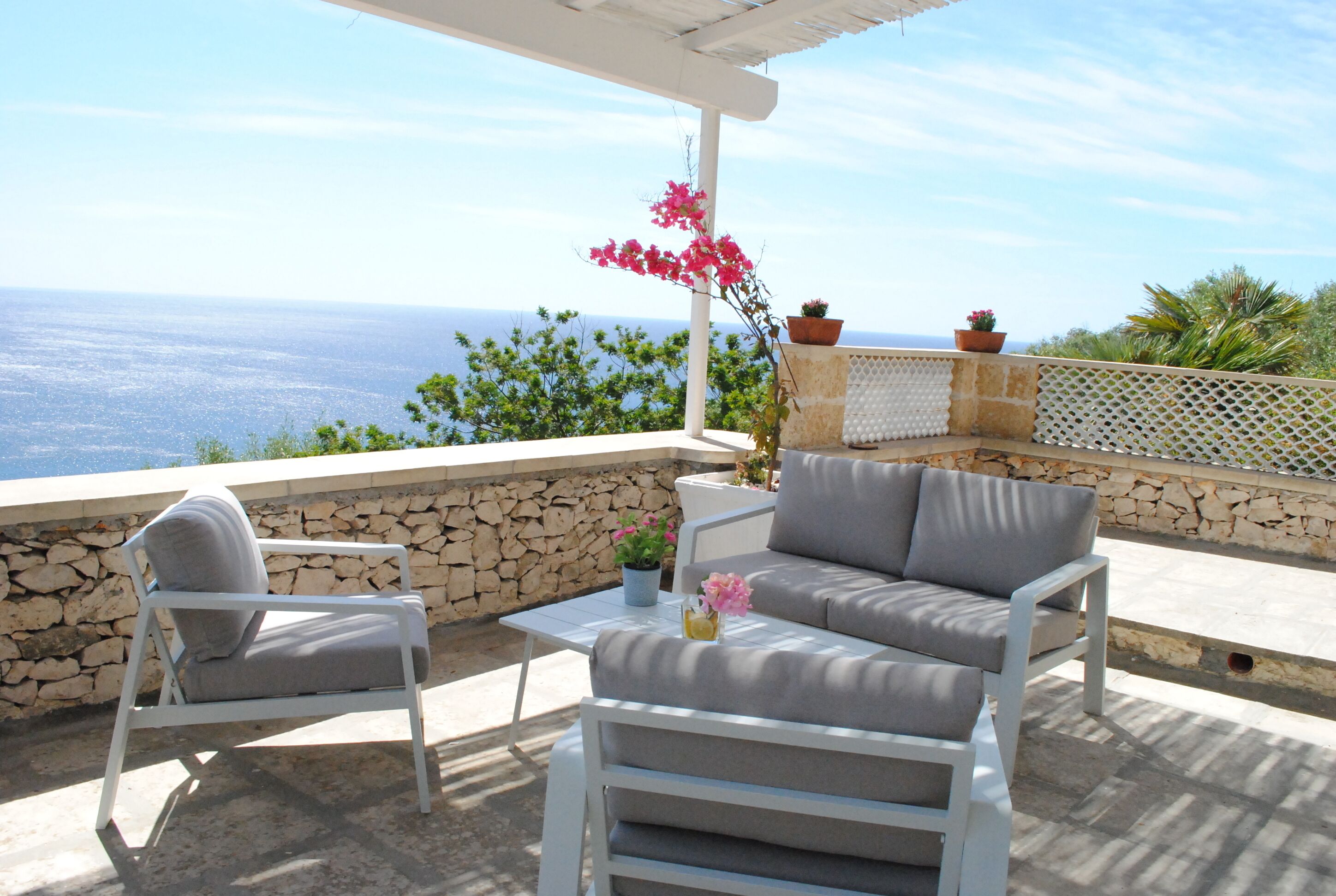 An outdoor seating area with a stone wall, white furniture, and a view of the sea.