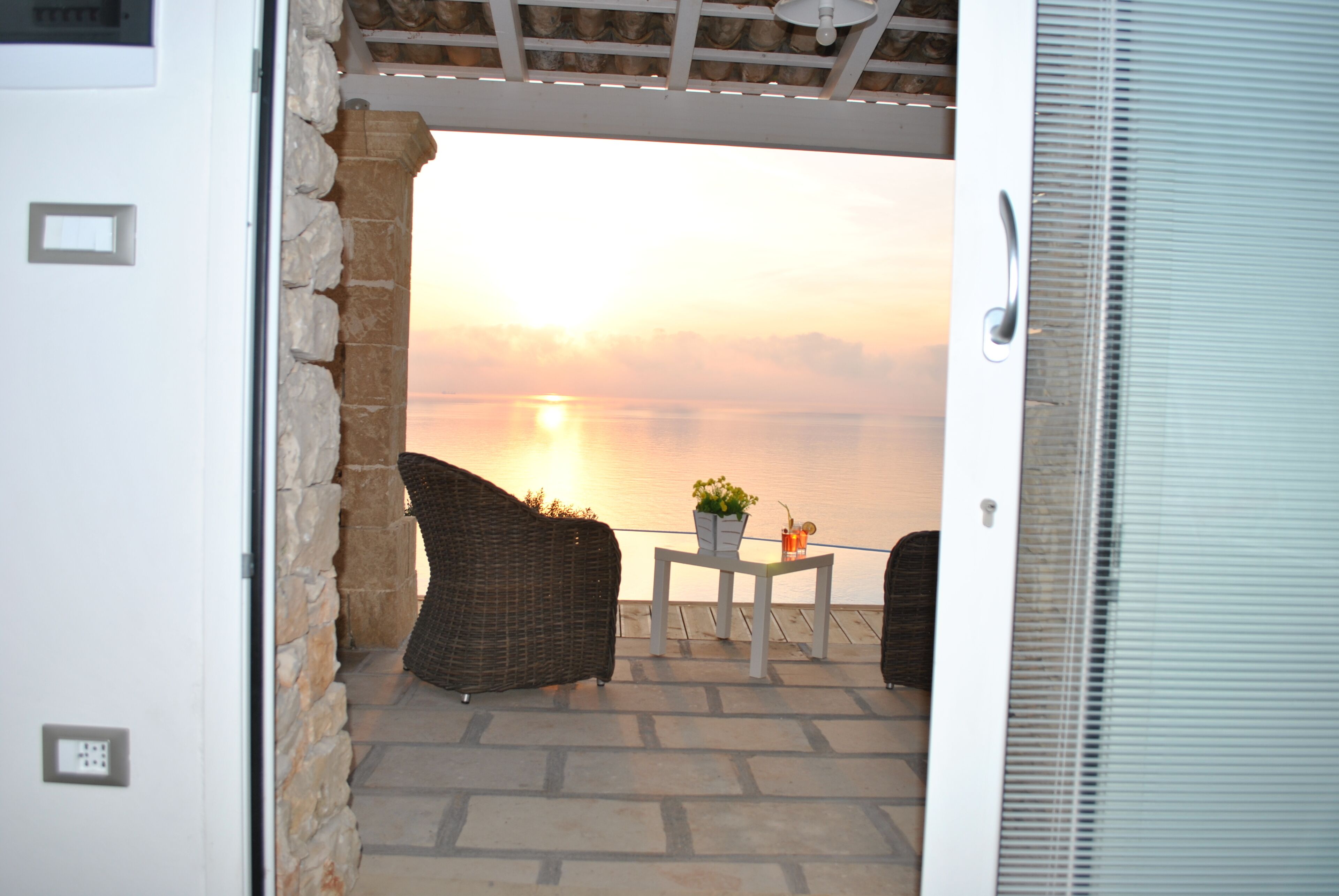 An open door leading to a patio with wicker furniture and a table with a plant and drink, overlooking a body of water at sunset.