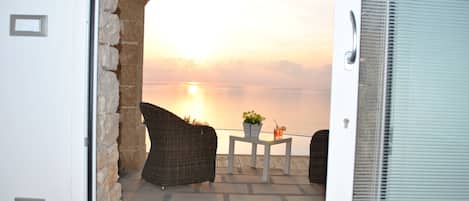 An open door leading to a patio with wicker furniture and a table with a plant and drink, overlooking a body of water at sunset.