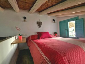 Ein Schlafzimmer mit Holzboden, einem Bett mit roter Bettdecke, einer herzförmigen Wanddekoration und einem Fenster mit blauer Tür.