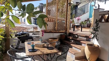 Een terras buiten met een houten tafel en stoelen, een barbecue en plantenpotten.