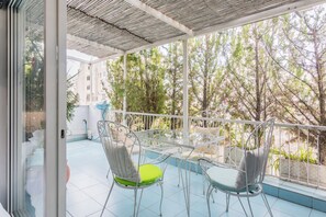 A balcony with a glass table and chairs, a metal railing, and a view of trees and buildings.
