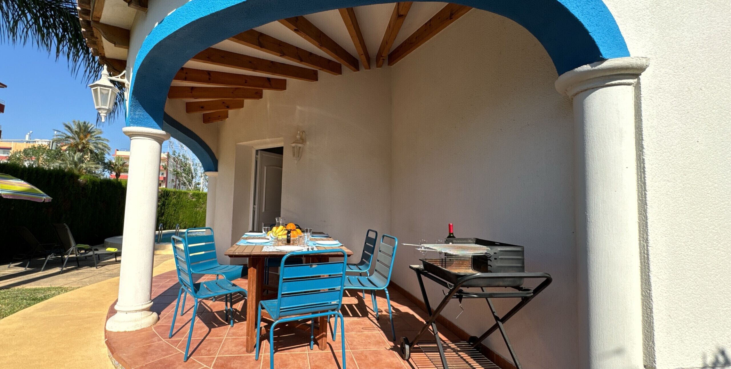 Une terrasse extérieure avec une table en bois et des chaises bleues, un barbecue et une piscine en arrière-plan.