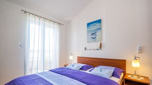 Une chambre à coucher comprenant un lit, deux tables de chevet, une commode, une lampe fixée au mur, une fenêtre avec des rideaux et un tableau accroché au mur.