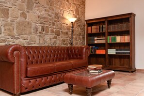 Um sofá de couro marrom com um banquinho combinando, uma estante de madeira cheia de livros e uma parede de pedra ao fundo.