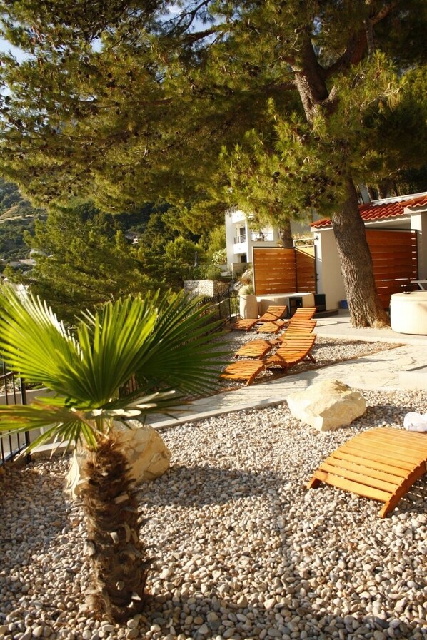 A patio area with wooden lounge chairs, a palm tree, and a rocky ground. - Apartment in Brela with Seaview, Terrace, Air condition, WIFI (3524-1) (Brela)