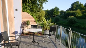 Una terraza con una mesa redonda y dos sillas, una planta en maceta y una barandilla con vista a un cuerpo de agua.