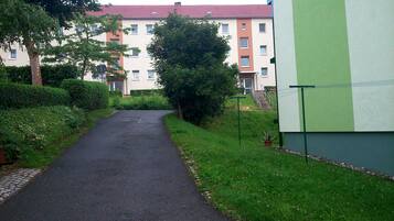 En smal, asfalteret sti, der fører til en bygning med røde tegltage, omgivet af grønt og en hæk.