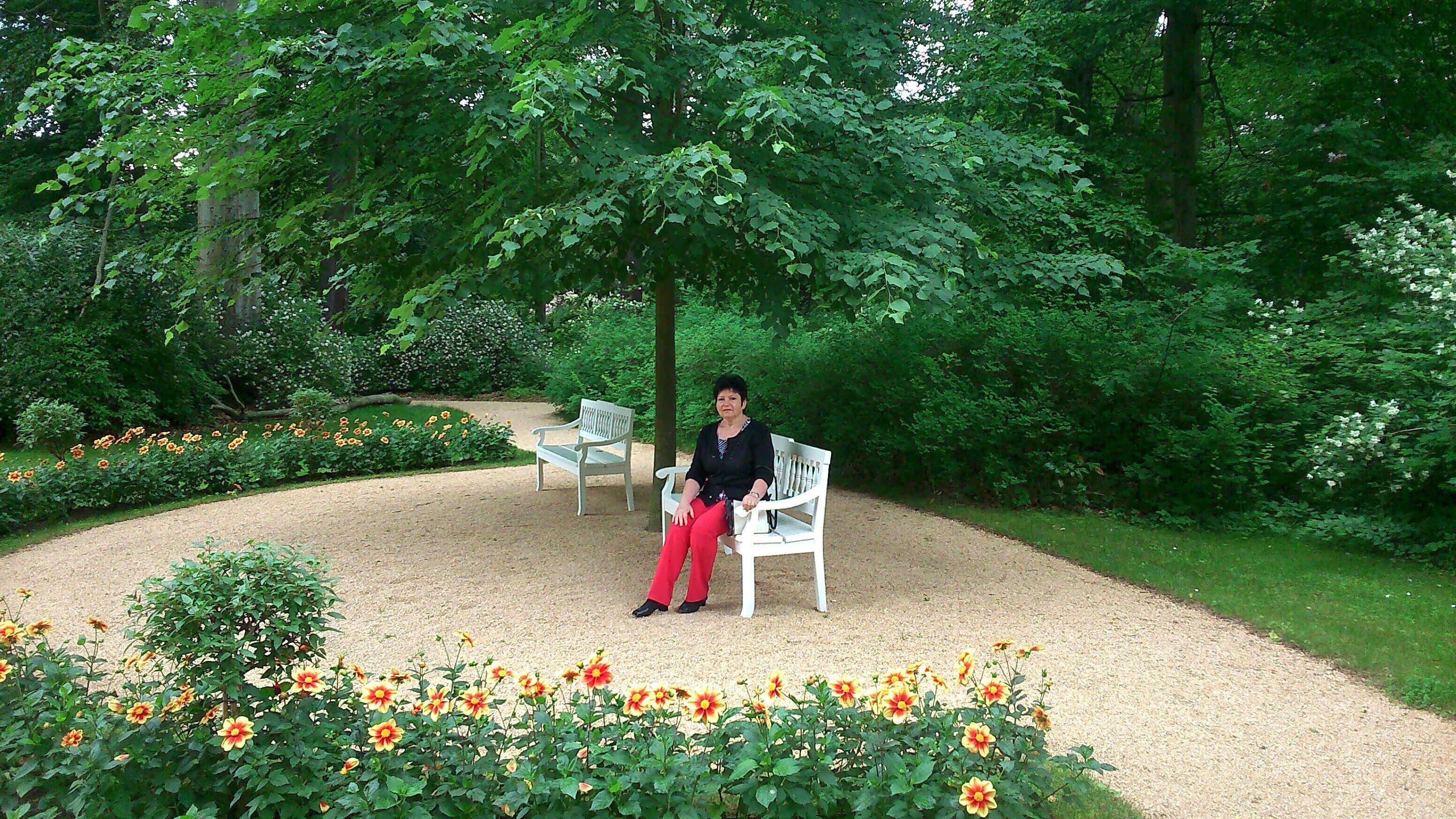 Eine Frau sitzt auf einer Bank in einem Garten mit Kiesweg, umgeben von viel Grün und Blumen.