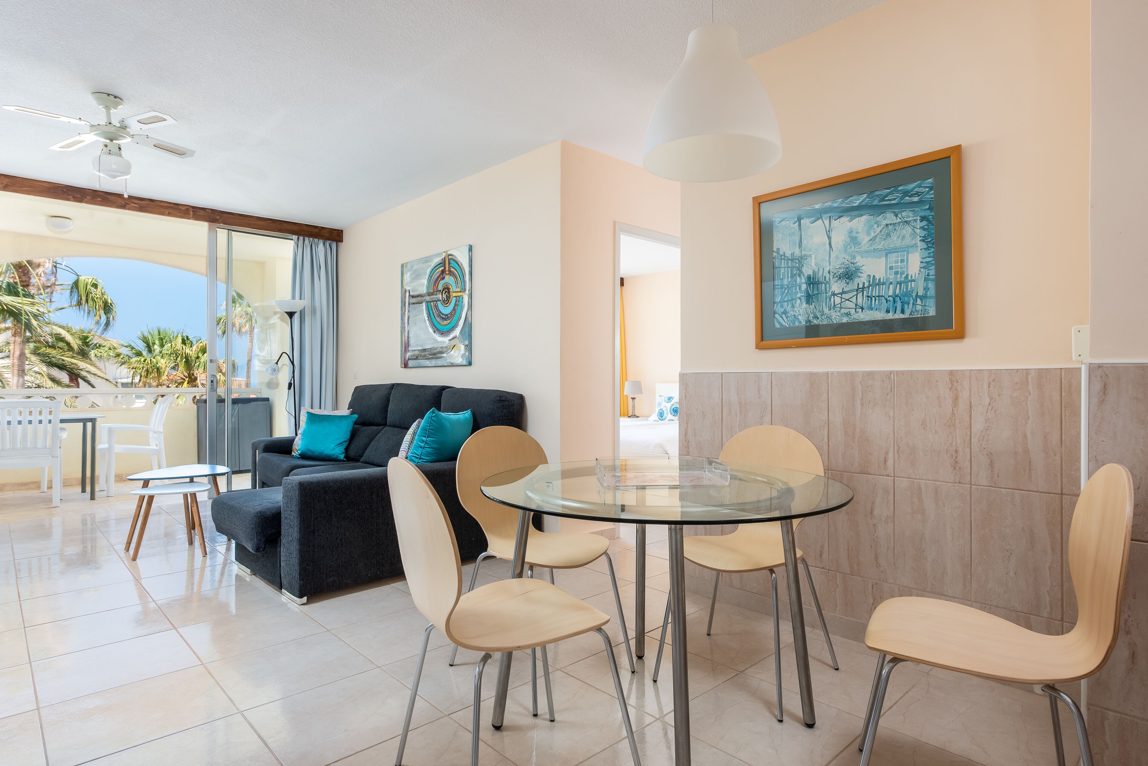Un salon moderne avec une table et des chaises en verre, un canapé noir agrémenté de coussins turquoise et un tableau accroché au mur.