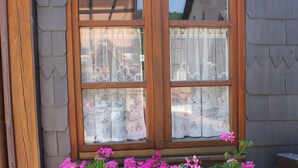 A window with a wooden frame and lace curtains, a brick wall, and a flower box with pink flowers. - Charming Cottage La Maisonnette Coquette (Oltingue)