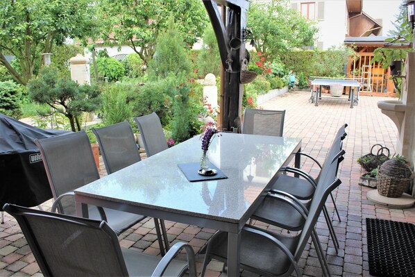 An outdoor patio with a table and chairs, surrounded by greenery and a brick floor.