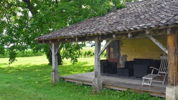 A rustic wooden structure with a tiled roof, a porch with wicker furniture, and a door leading inside. - quiet house on the banks of the Dordogne near Saint Emilion (Sainte-Terre)