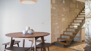 Ein moderner Essbereich mit einem runden Holztisch, vier Stühlen und einer besonderen Pendelleuchte. Im Hintergrund ist eine Treppe mit Holzboden und Metallgeländern zu sehen.