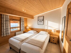 A wooden-paneled room with two beds, a window with curtains, a framed picture on the wall, and a nightstand with a lamp.