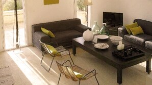 Un salon avec un canapé, une table basse et des fauteuils. Il y a des fenêtres et une lampe.