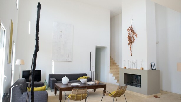 Un salon moderne avec un canapé gris, une table basse et deux fauteuils en osier. On y trouve une cheminée agrémentée d’objets décoratifs et un escalier menant à un autre niveau.