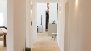 Un couloir doté d’une moquette de couleur claire, de murs blancs et de quelques meubles, dont une table et des chaises. Des luminaires décoratifs sont fixés aux murs, et un tableau encadré y est accroché.