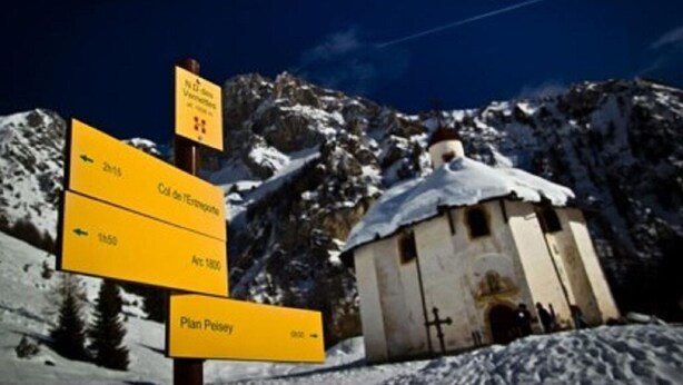Un cartel con indicaciones a varios lugares, un pequeño edificio con tejado nevado y un paisaje montañoso de fondo.