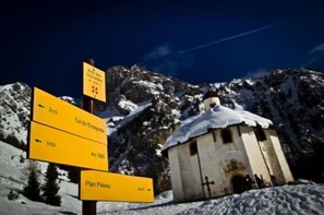 Un panneau indicateur indiquant les directions vers différents lieux, un petit bâtiment dont le toit est recouvert de neige, et un paysage montagneux en arrière-plan.
