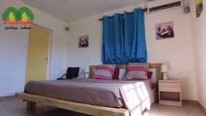 A bedroom with a bed, bedside tables, a nightstand, a window with blue curtains, and a wall-mounted air conditioner. - LA VILLA DES VOYAGEURS WITH POOL AND SPA FOR 4 TO 12 PEOPLE (SAINT FRANCOIS)