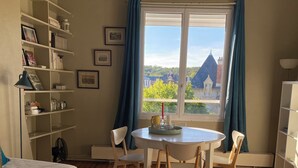 A room with a bed, a dining table with chairs, a small side table with a vase, and a window with curtains.