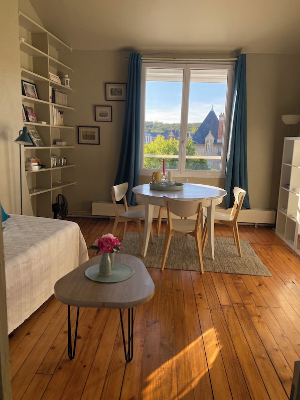 Une pièce comprenant un lit, une table à manger avec des chaises, une petite table d’appoint avec un vase, et une fenêtre avec des rideaux.