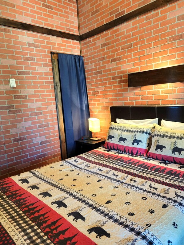 Una habitación con pared de ladrillo, una cama con ropa de cama estampada, una mesita de noche con lámpara y una ventana con cortina.