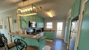 Una cocina con gabinetes color turquesa, encimera de granito, refrigerador de acero inoxidable e iluminación colgante.