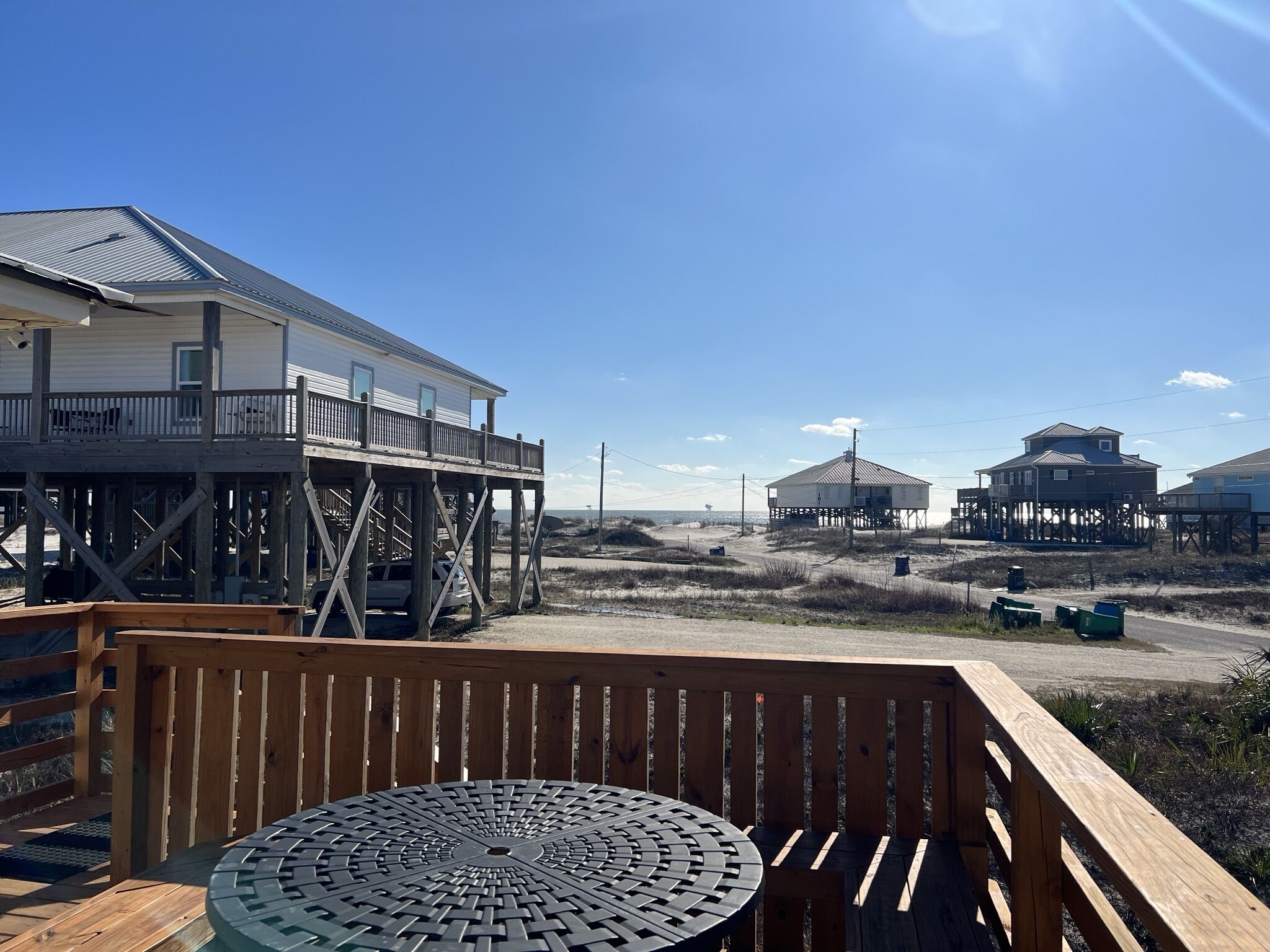 A wooden deck with a metal table, overlooking a beach with several beach houses and a clear sky.
