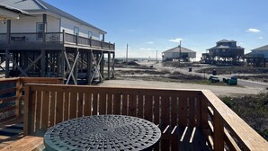 Una terraza de madera con una mesa metálica, con vistas a una playa con varias casas de playa y un cielo despejado.