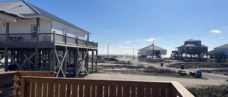 A wooden deck with a metal table, overlooking a beach with several beach houses and a clear sky.