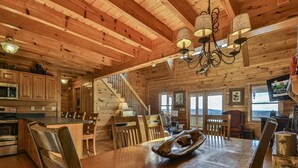 Interior de una cabaña de madera amplia, con una gran mesa de comedor, sillas de madera, una zona de cocina y una escalera que sube a otro nivel.