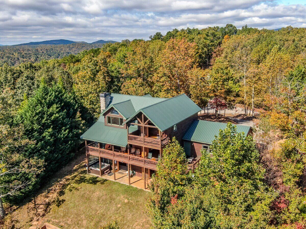 A large, two-story log cabin with a green roof, surrounded by dense forest and hills in the background.