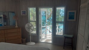 Een slaapkamer met een bed, een dressoir, een ventilator en grote ramen die uitkomen op een balkon.