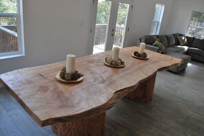 A large wooden dining table with three candles and decorative items in a modern living room with a gray sofa and a view of a deck through glass doors.