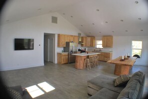 A spacious living area with a large wooden dining table, a built-in kitchen with wooden cabinets, and a flat-screen TV mounted on the wall.