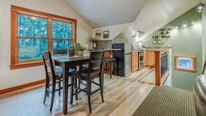 A kitchen with a dining area, wooden flooring, a ceiling fan, and a window with a view of trees. - Close-Country Retreat (Eugene)