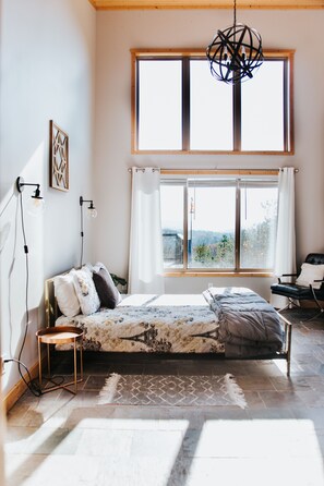 A bedroom with a large window, a bed with a patterned bedspread, a standing lamp, and a chair.