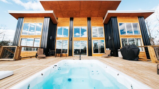 A modern wooden cabin with a hot tub on a deck, large windows, and a clear sky.