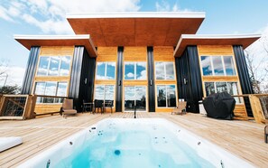 A modern wooden cabin with a hot tub on a deck, large windows, and a clear sky.