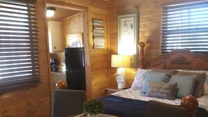 En mysig stuga med en inredning som inkluderar ett matbord i trä, två stolar, en säng med kuddar och ett litet köksutrymme.