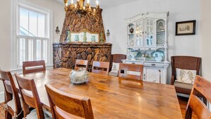 A dining room with a large wooden table, six chairs, a stone fireplace, and a white cabinet with glass doors.