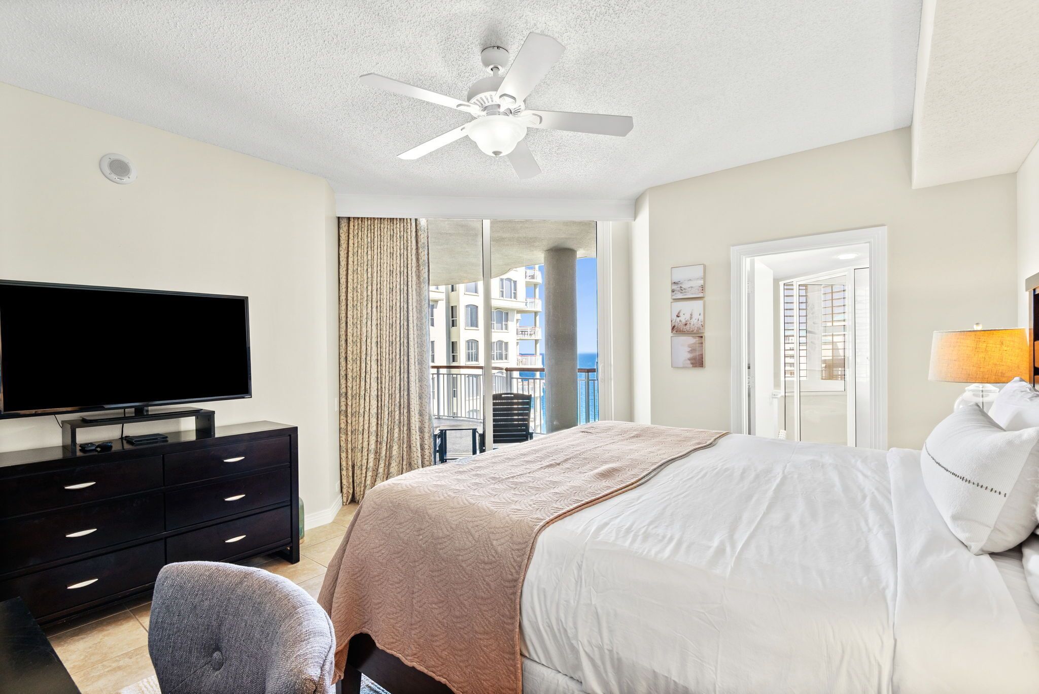 A hotel room with a bed, a TV, a ceiling fan, a window with a view of buildings, and a door.