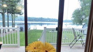 Uitzicht vanuit een kamer met een groot raam op een terras met een witte reling, een barbecue en een boot op de achtergrond.