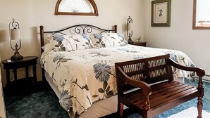 Een slaapkamer met een bed, twee nachtkastjes, een bankje, een wandventilator en een lijst met een afbeelding.