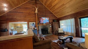 Un interior de cabaña acogedor, con techo de madera, chimenea de piedra, televisor y comedor.