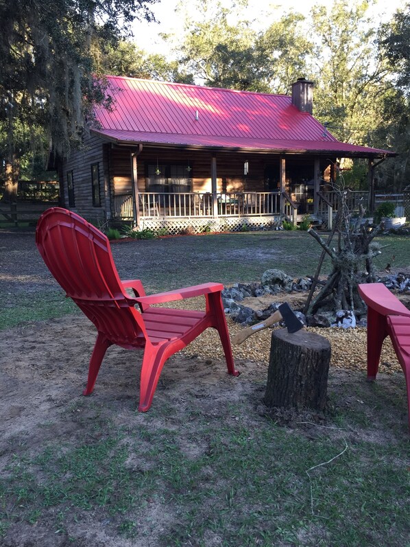 A wooden cabin with a red roof, a covered porch, and a fire pit area with two red Adirondack chairs.