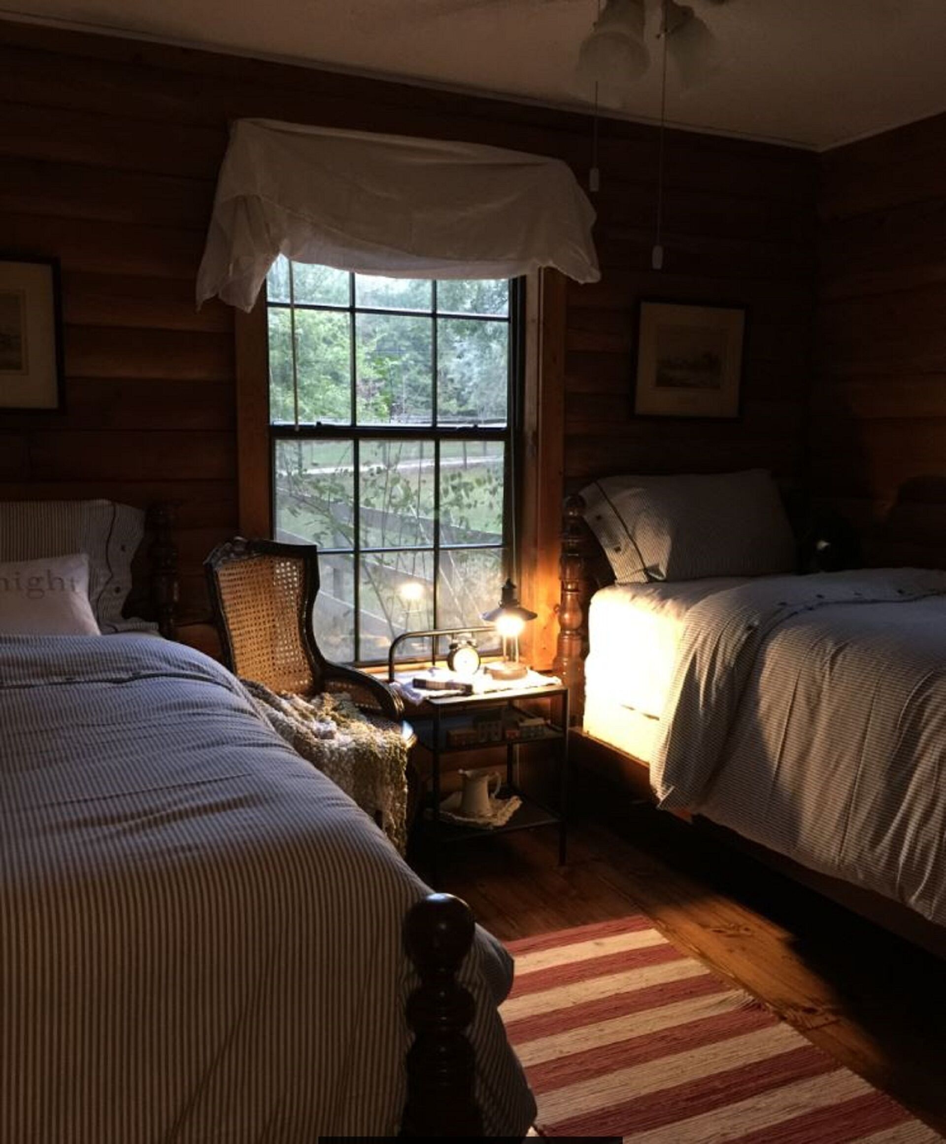 A cozy cabin-style room with two beds, a wooden floor, a window with a view of greenery, and a small table with a lamp.