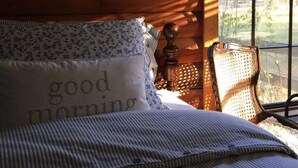 Una cama con ropa de cama a rayas y una almohada con el texto "good morning", una silla de mimbre y una ventana con vistas a árboles.